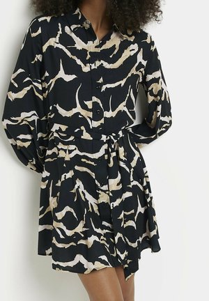 Femme portant une robe à boutons noire et beige à motif abstrait, avec des manches longues bouffantes et une ceinture nouée à la taille.