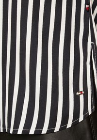 Gestreiftes Hemd mit vertikalen schwarz-weißen Linien, ausgestattet mit einem kleinen, farbigen Logo nahe dem Saum. Strukturierter Stoff mit taillierter Passform.