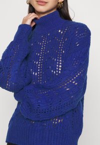 Pletený svetr v zářivě modré barvě, s vysokým límcem, texturovanými průsvitnými vzory a mírně oversized rukávy. Měkký, pohodlný materiál.