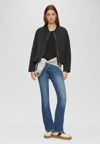 Giacca bomber nera con zip argento, abbinata a un top nero aderente, una felpa grigia chiara legata in vita e jeans svasati blu.