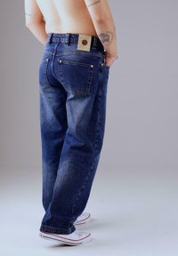 Person, der lockere dunkelblaue Jeans und weiße Turnschuhe trägt, mit den Händen in den Taschen steht und tätowierte Arme vor einem neutralen Hintergrund zeigt.