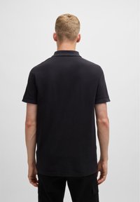 Homme avec les cheveux blonds courts portant un polo noir à manches courtes uni, se tenant face à un fond gris clair.