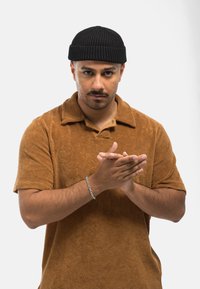 Polo shirt in terry cloth marrone a maniche corte con colletto pieghevole, abbinato a un berretto di maglia nero; il modello applaude, indossando un bracciale d'argento.