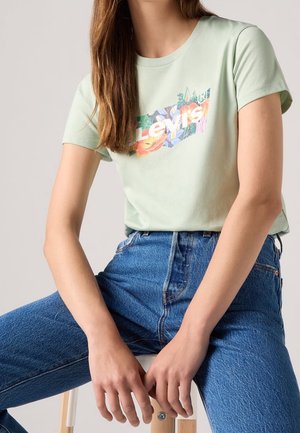 Osoba ubrana w jasnorozowy kwiatowy t-shirt z logo Levi's i niebieskie dżinsy, siedząca na drewnianym stołku z rękami spoczywającymi z przodu.