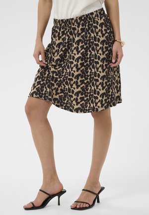 Žena nosiaca sukňu s leopardím vzorom po kolená, biely top, čierne sandále na podpätku a zlatý náramok stojaca na svetlom pozadí.