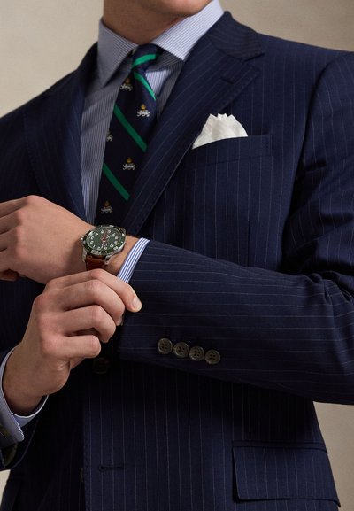 Σκούρο μπλε κοστούμι με λωρίδες και ανοιχτό μπλε πουκάμισο με ρίγες. Διαθέτει γραμμωτή πράσινη γραβάτα και ασημένιο-μαύρο ρολόι με καφέ δερμάτινο λουράκι.