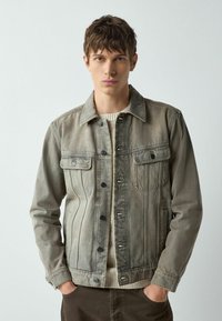 Džinsa jaka klusās pelēkās nokrāsās ar divām krūšu kabatām, pogu aizdari un smalku izbalējumu, vilkta virs krēmkrāsas džempera.