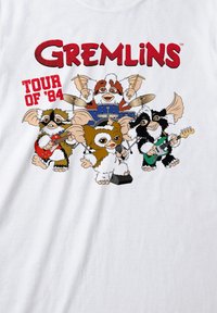 Weißes T-Shirt mit cartoonhaften Gremlins-Charakteren, die Instrumente spielen. Der Text lautet "GREMLINS TOUR OF '84" in Rot, mit bunten Akzenten.
