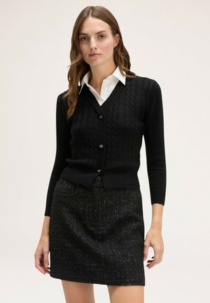 Cárdigan negro de punto con un escote en V y un diseño ajustado, que presenta un patrón de trenzas texturizadas y botones dorados, combinado con una falda negra texturizada.