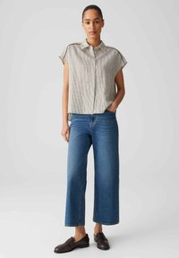Camicia a maniche corte con colletto a righe orizzontali nei colori beige e marrone scuro, abbinata a jeans in denim blu a gamba larga e mocassini neri.