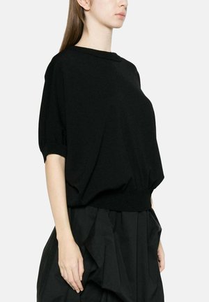 Jeune femme portant un pull ample noir à manches courtes et une jupe noire avec un détail de tissu froncé, vue de profil.