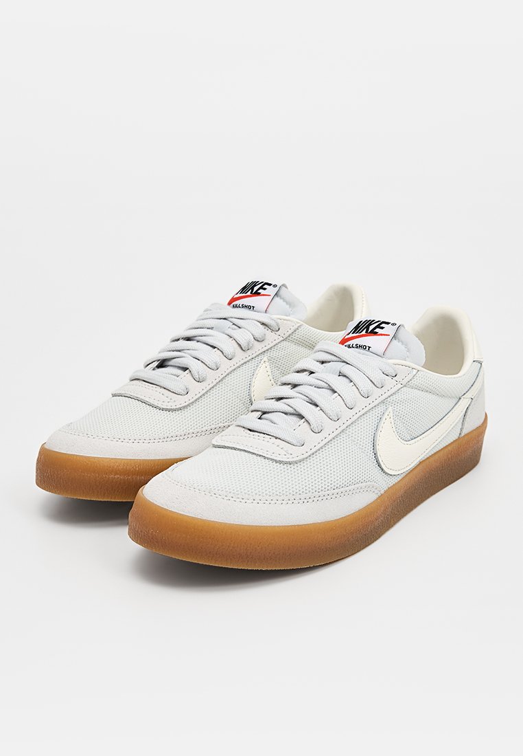 Zapatillas de malla gris claro con acentos en crema, cordones planos y suelas de goma. Presentan un logo de Nike y un collar acolchado.