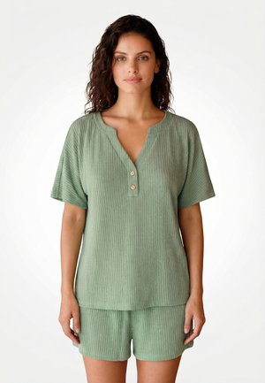 Vrouw met krullend haar, gekleed in een lichtgroen geribd topje met korte mouwen en knopen, en bijpassende korte broek, staand tegen een effen witte achtergrond.