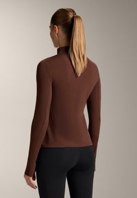 Top lung cu mânecă lungă, de culoare maro, cu guler înalt și design ajustat, realizat dintr-un material neted, asortat cu colanți negri.