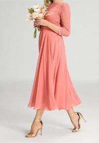 Robe corail avec manches trois-quarts en dentelle, jupe fluide, et sandales à talons hauts dorées. Le mannequin tient un bouquet de fleurs.