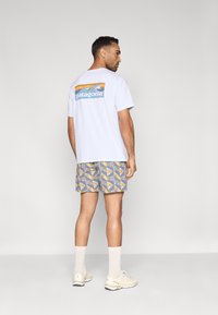 Λευκό t-shirt με μπλε και πορτοκαλί λογότυπο Patagonia στην πλάτη, σε συνδυασμό με σορτς με σχέδιο που απεικονίζει κίτρινες κυμάτιες σε μπλε φόντο.