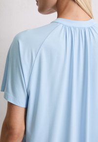 Lichtblauw, kortärmige top met een ribbelkraag en geplooide details aan de achterkant, gemaakt van een zachte, gladde stof.