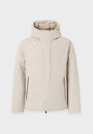 Homme portant une veste à capuche beige clair, une chemise blanche, un pantalon cargo beige et des baskets marron foncé, debout devant un fond uni.