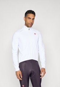 Weiße Radjacke mit hohem Kragen und durchgehendem Reißverschluss, mit rotem Logo auf der Brust. Getragen mit dunklen Sporttights. Glatte Textur.