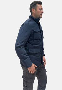 Giacca blu navy di peso medio con multiple tasche applicate, chiusure a scatto e collo alto. Pantaloni cargo beige completano l'outfit.