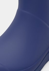Cizma din cauciuc de culoare bleumarin, cu o textură netedă, formă rotunjită și o ușoară curbă la gleznă. Are o finisare lucioasă.