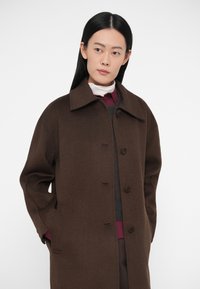 Cappotto di lana marrone con ampio colletto, bottoni frontali e tasche laterali. Indossato sopra un maglione grigio e una camicetta bordeaux. Texture liscia e vestibilità rilassata.