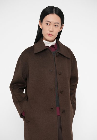 Manteau en laine marron avec un large col, des boutons sur le devant et des poches sur les côtés. Porté sur un pull gris et un chemisier bordeaux. Texture lisse et coupe décontractée.