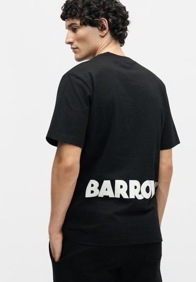 Άνδρας με σγουρά μαλλιά που φοράει μαύρο μπλουζάκι με τη λέξη «BARROW» τυπωμένη με μεγάλα λευκά γράμματα στο κάτω μέρος της πλάτης.