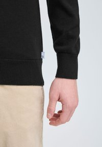 Maglione nero a maglia con polsini a costine, con un'etichetta visibile di lato. Una mano è appoggiata accanto a pantaloni in velluto a coste beige.