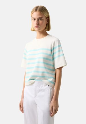 Blonde woman standing wearing white short-sleeve shirt with light blue horizontal stripes and white pants against plain background.