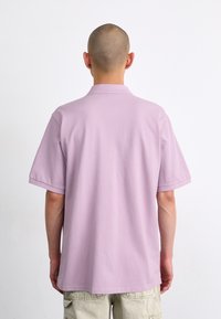 Polo lilas à manches courtes, avec un col classique et une texture de tissu lisse. La vue de dos montre une coupe décontractée et sans détails visibles.