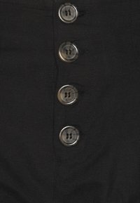 Schwarzer Stoff mit einer markanten vertikalen Reihe von fünf klaren Knöpfen. Die Knöpfe verfügen über zwei Knopflöcher und ein glänzendes Finish.