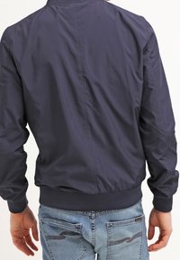 Mörkblå bomberjacka med en slät yta, ribbad mudd och fåll, samt en ren design. Bärs över ljusblå jeans med detaljer.