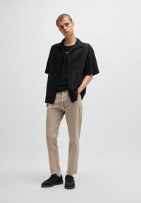 Camicetta nera a maniche corte con motivo floreale, abbinata a jeans slim-fit beige e sneakers nere, con una semplice t-shirt nera sotto.