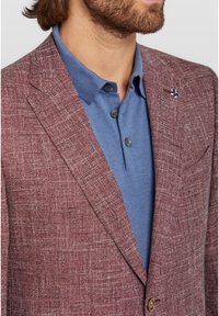 Blazer borgogna con texture e un motivo sottile, abbinato a un polo blu con tre bottoni. Include una spilla decorativa per il risvolto.