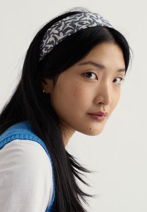 Ung kvinde med langt sort hår iført mønstret pandebånd og blå ærmeløs sweater over hvid skjorte, ser til siden mod en ensfarvet baggrund.