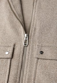 Beige Fischgrätenstoffjacke mit silbernem Reißverschluss und zwei Klappentaschen, die mit silbernen Druckknöpfen gesichert sind.