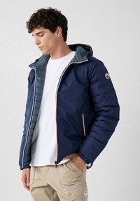 Hombre con chaqueta con capucha azul marino sobre camisa blanca y pantalones cargo beige, de pie con una mano en el bolsillo, mirando hacia abajo a la izquierda.