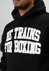 Zwarte hoodie met gedurfde, witte, verweerde tekst "HE TRAINS FOR BOXING" aan de voorkant. Materiaal lijkt zacht met een gestructureerde afwerking.
