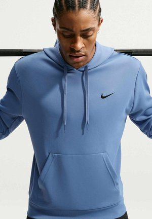 Homme en sweat à capuche bleu Nike reposant une barre noire sur ses épaules, regardant vers le bas avec les cheveux tressés.