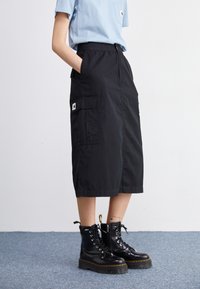 Black cargo skirt with an elastic waistband, featuring two large side pockets and a logo tag. Paired with black lace-up boots.