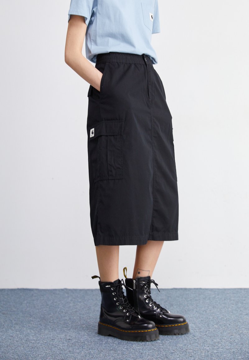 Black cargo skirt with an elastic waistband, featuring two large side pockets and a logo tag. Paired with black lace-up boots.