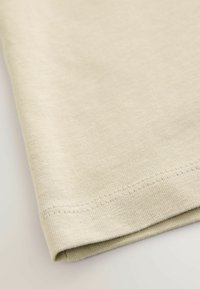Beige bomuldsstof tæt på med en glat tekstur. Kanten har en foldet kant og pæne sømme.