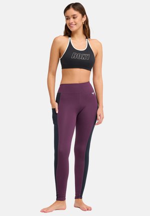 Jeune femme souriante, debout pieds nus, portant un soutien-gorge de sport noir avec le logo ROXY et des leggings violets avec des panneaux latéraux noirs et une poche.
