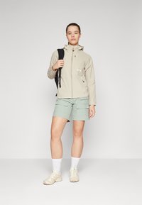 Veste imperméable beige clair avec fermeture éclair à l'avant, associée à un short cargo vert menthe. Le modèle porte des baskets claires et des chaussettes blanches.
