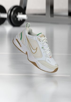 Baskets Nike blanches et beiges avec des accents verts flottant au-dessus d'une surface réfléchissante avec des poids de salle de sport flous en arrière-plan.