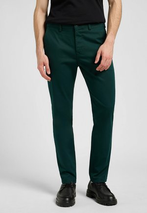 Green trousers made of smooth fabric, featuring a slim fit, front pockets, and a zip fly. Worn with black shoes.