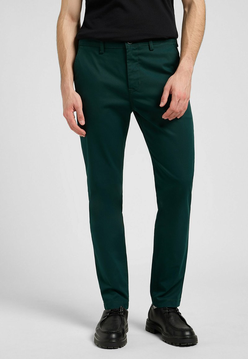 Grüne Hose aus glattem Stoff, mit slim fit, Fronttaschen und Reißverschluss. Getragen mit schwarzen Schuhen.