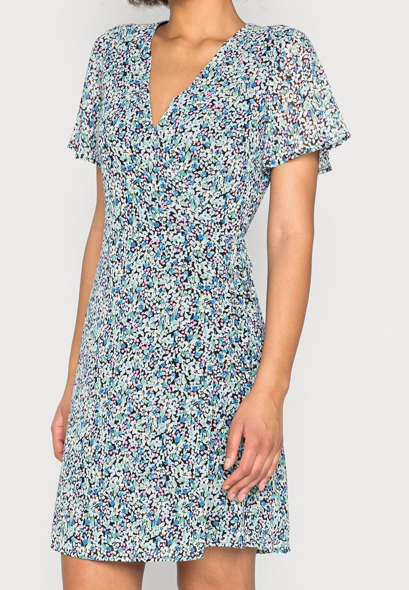 Femme portant une robe florale à manches courtes et col en V avec des fleurs bleues, roses et blanches sur fond noir.