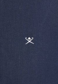 Biała haftowana figura skrzyżowanych pastorałów z dzwonkiem powyżej na ciemnoniebieskiej teksturowanej tkaninie.